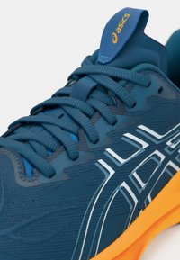 Blauwe atletische schoen met een gestructureerde bovenkant, contrasterend wit logo en oranje zool. De veters zijn dik en een trekhaak is zichtbaar op de tong.