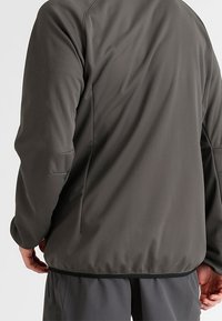 Mann in einer dunkelgrauen langärmeligen Jacke und passenden Hosen, von hinten gezeigt, mit entspannten Händen an den Seiten.