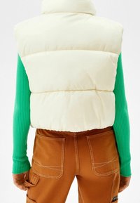 Cream puffer vest with a high collar, side seams, and a smooth exterior, worn over a green ribbed long-sleeve top and brown pants.