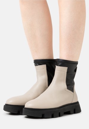 Stivaletti in pelle beige a caviglia con un alto elastico nero, suola in gomma texture e cuciture evidenti lungo le giunture.