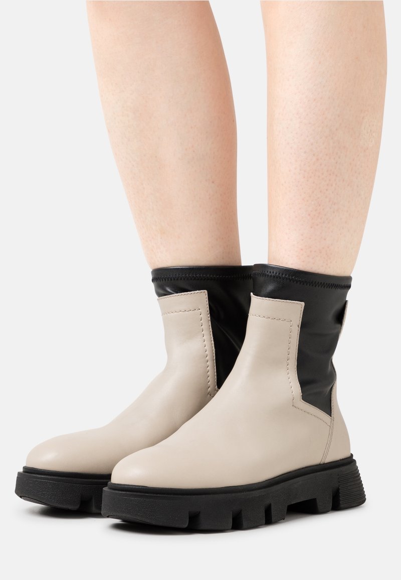 Botas de tobillo de cuero beige con parte superior elástica negra, suela de goma texturizada y costuras prominentes a lo largo de las costuras.
