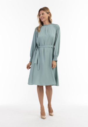 O rochie verde mentă, până la genunchi, cu mâneci lungi, un detaliu cu umeri strânși și o curea din același material la talie. Țesătura are o textură netedă.