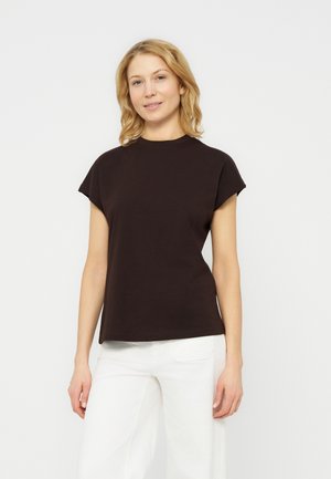 Kobieta o blond włosach, ubrana w czarną bluzkę z krótkim rękawem i białe spodnie, stojąca na jednolitym jasnym tle.