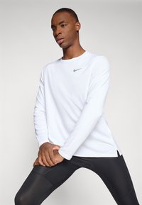 White long-sleeve sports shirt with a round neckline and a gray Nike logo. Paired with black fitted leggings, emphasizing athletic fit.