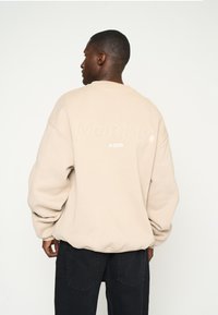 Beige Crewneck-Sweatshirt mit oversized Passform, mit einer strukturierten Logostickerei "Multiply" und dem Jahr "2015" auf dem Rücken.