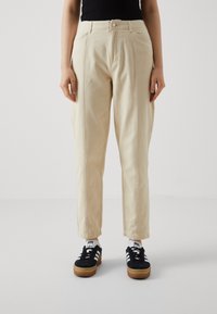 Pantalon beige taille haute avec plis à l'avant et coupe décontractée, en tissu de coton doux. Porté avec des baskets noires ornées de bandes blanches.