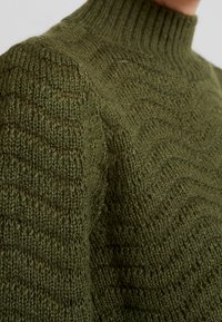 Maglione lavorato a maglia di colore verde scuro con colletto alto a costine, caratterizzato da un motivo a onde testurizzato su tutta la stoffa. Tessuto morbido e caldo.