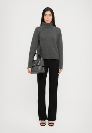 Donna che indossa un maglione grigio a collo alto, pantaloni neri a zampa d'elefante, sandali neri, e porta una borsa nera sulla spalla su sfondo bianco.