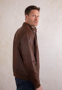 Man wearing a brown leather jacket and jeans, standing sideways, looking over his shoulder against a plain light background.