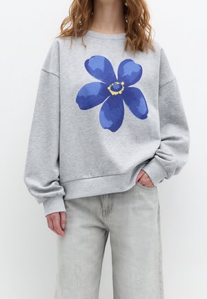 Femme portant un sweat-shirt gris clair avec un grand motif fleur bleu et un pantalon gris clair, debout avec une main dans la poche.