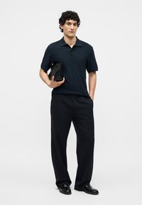 Mann in dunklem Poloshirt und weitem Hosen hält eine schwarze gewebte Clutch, steht mit einer Hand in der Tasche vor weißem Hintergrund.