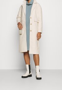 Cappotto di lana color crema con collo, due tasche frontali e bottoni dorati. Indossato sopra un vestito turchese, abbinato a stivaletti bianchi e neri.