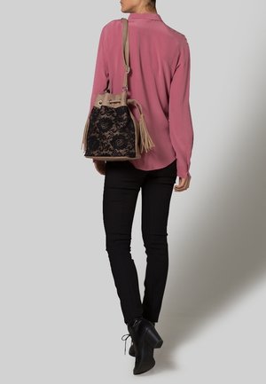 Femme portant un chemisier rose, un pantalon noir et des chaussures à talons noires, portant sur l'épaule un sac seau beige et noir à motif de dentelle avec des pompons.