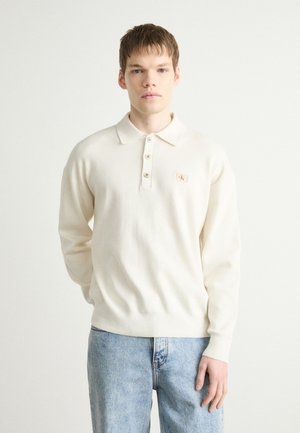 Maglione polo color crema con una consistenza morbida, tre bottoni e una piccola etichetta con logo. Abbinato a jeans denim blu chiaro.