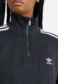 Juodas zip-up džemperis, pagamintas iš medvilnės mišinio, su baltais pečių raižais ir mažuoju baltu Adidas logotipu kairėje krūtinės pusėje.