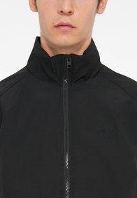 Black zip-up jacket with high collar, made from smooth, lightweight material. Features a subtle logo and no visible patterns or hardware.