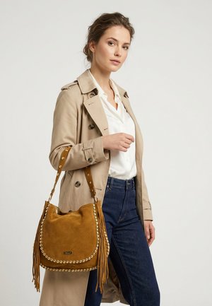 Kobieta w beżowym płaszczu trencz i białej koszuli trzyma brązową zamszową torebkę z złotymi ćwiekami i frędzlami, nosi ciemnoniebieskie dżinsy na jednolitym tle.