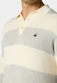 Polo tričko s proužky v krémové a světle šedé barvě; má límec, tři knoflíky a vyšitým logem na prsou. Měkká pletená struktura.