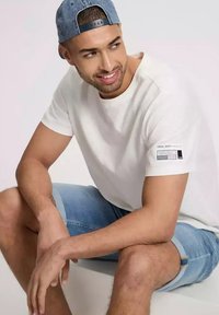 Biała bawełniana koszulka z krótkim rękawem, z małą metką na lewym rękawie i luźnym krojem. W zestawie z niebieskimi szortami dżinsowymi.