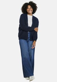 Jūras spēku zila adīta kardigāna josta ar brīvu piegriezumu, garām piedurknēm un V-veida dekoltē, kombinēta ar gaiši pelēku T-kreklu un plašām zila džinsu biksēm.