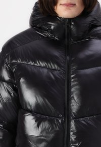 Sort pufferjakke med en blank finish, høj krave og lynlås foran. Har quilte-sektioner og en tætsiddende design for varme.