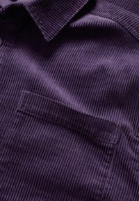 Violeta korduroja audums ar vertikālām rieviņām, kas ietver apkakli un uzšūtu kabatu. Teksturēta virsma sniedz dziļumu un dimensiju.