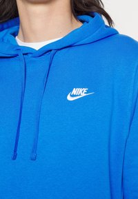 Modrá mikina Nike s bílým vyšitým logem na hrudi, nošena přes bílou košili; mikina má šňůrku se viditelnými smyčkami.