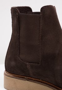 Botín de ante marrón con paneles laterales elásticos, una costura y una suela de goma de color claro. Vista en primer plano del tacón y la textura.