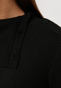 Pull noir à côtes avec un col haut et deux boutons décoratifs à l'épaule. Texture douce avec une coupe ajustée.