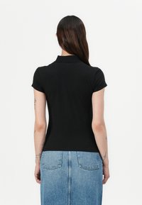 Zwarte polo-shirt met korte mouwen, aansluitend ontwerp. Achteraanzicht toont een gladde textuur en kraag. Gecombineerd met een spijkerrok.