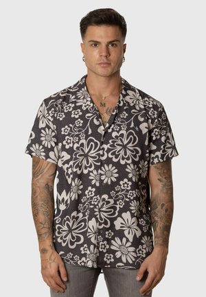 Hombre con cabello corto y oscuro y tatuajes, que lleva una camisa de botones de manga corta con estampado floral en blanco y negro y pantalones grises.