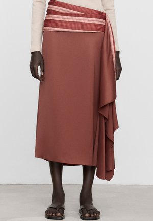 Jupe midi marron avec un détail drapé sur le côté, associée à un haut à manches longues crème et des sandales noires à bout ouvert sur une personne à la peau foncée debout.
