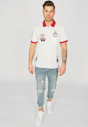 Hvid polo skjorte med rød krave og detaljer, der har broderede lapper og en brystlomme. Båret med lys blå slidte jeans og hvide sneakers.