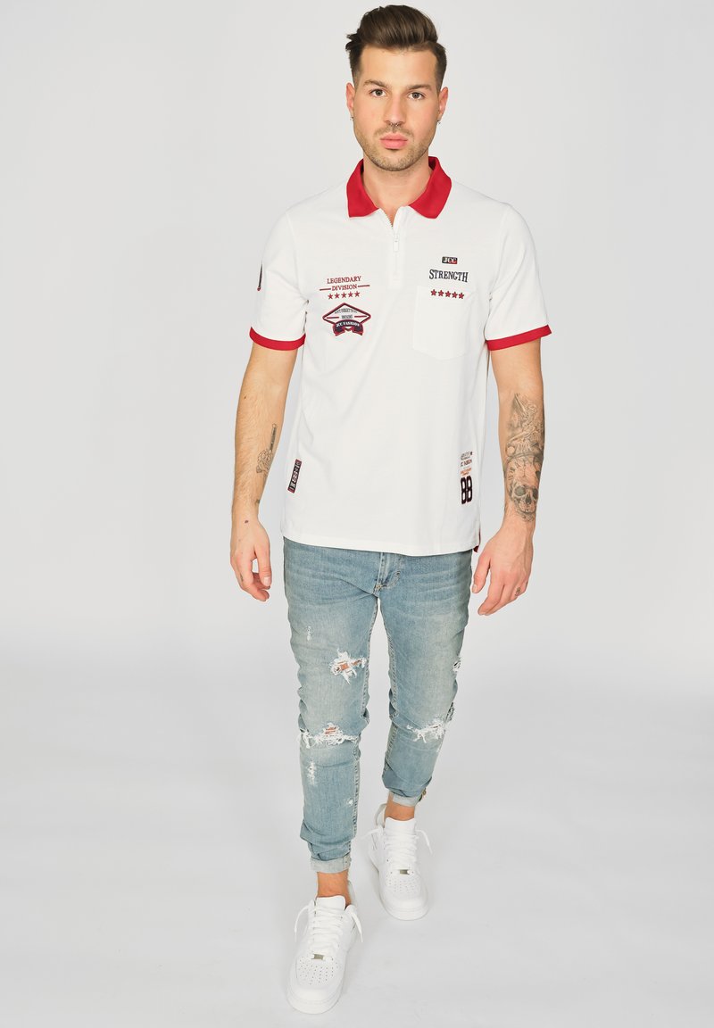 Vit polo med röd krage och detaljer, utrustad med broderade lappar och en bröstficka. Bärs med ljusblå slitna jeans och vita sneakers.