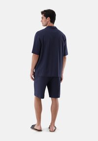 Marineblaues Kurzarmhemd und passende Shorts aus strukturiertem Material. Das Outfit hat einen Kragen und einen lässigen Schnitt. Sandalen runden den Look ab.