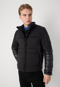 Black puffer jacket with a textured matte finish, zippered side pockets, and a detachable hood. Features padded design and adjustable cuffs.