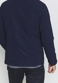 Giacca blu navy con finitura testurizzata, caratterizzata da una linea dritta, maniche lunghe e orlo a bandella. La vista dal retro mostra dettagli minimi.