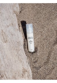 Flacon de Susanne Kaufmann Ectoin Barrier Repair Moisturiser SPF 50 posé sur un sol sablonneux à côté d'un morceau de bois flotté usé par le temps.
