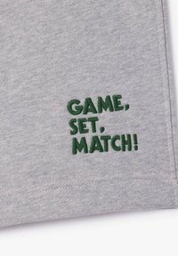 Tissu gris avec texte brodé en vert "MATCH, SET, GAME !" près du bord, suggérant un thème sportif ou de tennis.