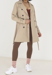 Beżowy trencz z podwójnymi guzikami, naramiennikami i wiązaniem w talii. Nosi się go na brązowe prążkowane legginsy oraz białe sneakersy.