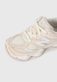 Sapatilha New Balance 9060 em bege e branco com parte superior em malha e camurça, atacadores brancos e sola robusta sobre fundo branco.