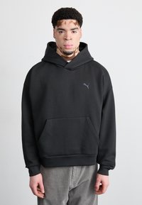 Sort hætte sweatshirt med en frontlomme og et lille gråt Puma-logo på brystet. Stoffet ser glat og tykt ud og har en afslappet pasform.