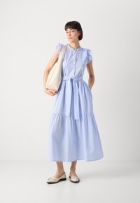 Gestreiftes Kleid in Blau und Weiß mit Rüschensäumen, Knopfverschluss vorne, Taillengürtel und gestuftem Rock; kombiniert mit einer beigen Schultertasche und flachen Schuhen.