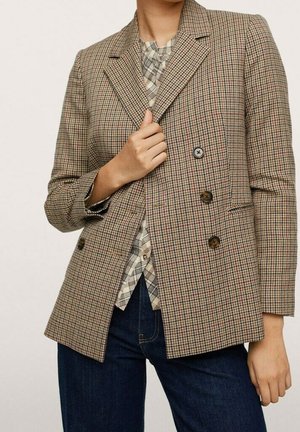 Person trägt einen braunen karierten zweireihigen Blazer über einem beigen karierten Hemd und dunkelblauen Jeans, mit einer Hand, die den Blazer-Revers hält.