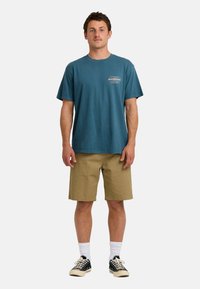 Turkusowy bawełniany t-shirt z okrągłym dekoltem, krótkimi rękawami i logo z przodu; zestawiony z khaki szortami i czarnymi sneakersami z płótna.