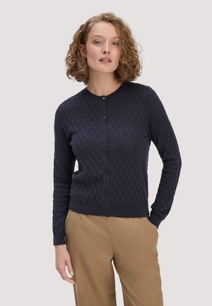 Frau mit lockigem Haar trägt eine marineblaue, strukturierte, geknöpfte Strickjacke und beigen Hosen, steht mit einer Hand in der Tasche.