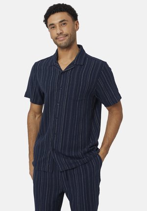 Mężczyzna ubrany w granatową koszulę na krótki rękaw z guzikami oraz pasujące spodnie z cienkimi białymi pionowymi prążkami, stojący z jedną ręką w kieszeni.