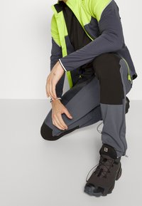 Tenue sportive comprenant une veste grise avec des accents vert lime, associée à un pantalon noir. Les détails notables incluent des zippers et un tissu texturé.