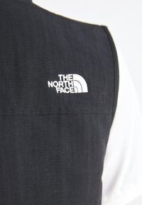 Czarna kamizelka z prążkowanym wzorem, z białym haftowanym logiem "The North Face" na górnej lewej stronie. Prosty design, bez widocznych akcesoriów.