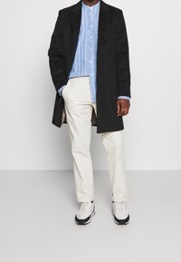 Uomo che indossa un cappotto nero, una camicia a righe blu, pantaloni color crema e sneakers bianche, in piedi con una mano in tasca.